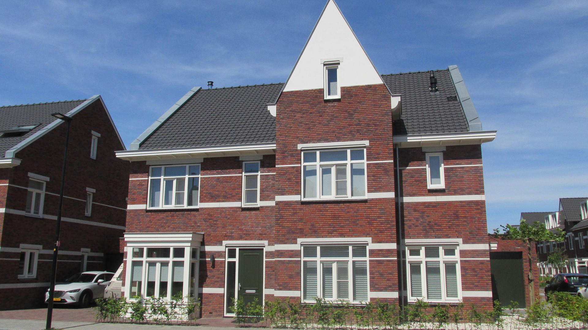 Moderne rode bakstenen woning met witte kozijnen en een driehoekig dak, gelegen in een woonwijk onder een blauwe lucht.