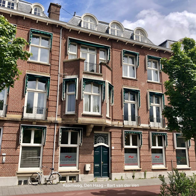 Bakstenen gebouw met groene luiken, het adres 'Raamweg, Den Haag' onderaan zichtbaar, een fiets staat links van de deur.