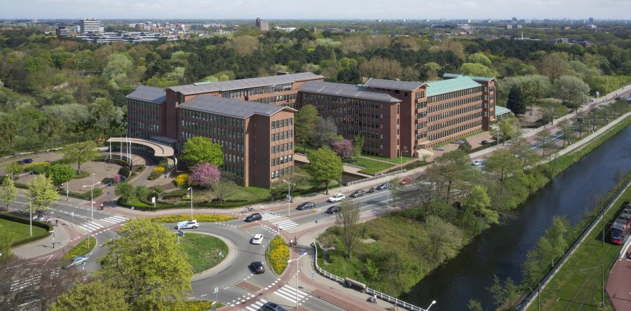 Luchtfoto van een modern kantoorgebouw omringd door groen, met een drukke weg en een ronde rotonde ervoor.