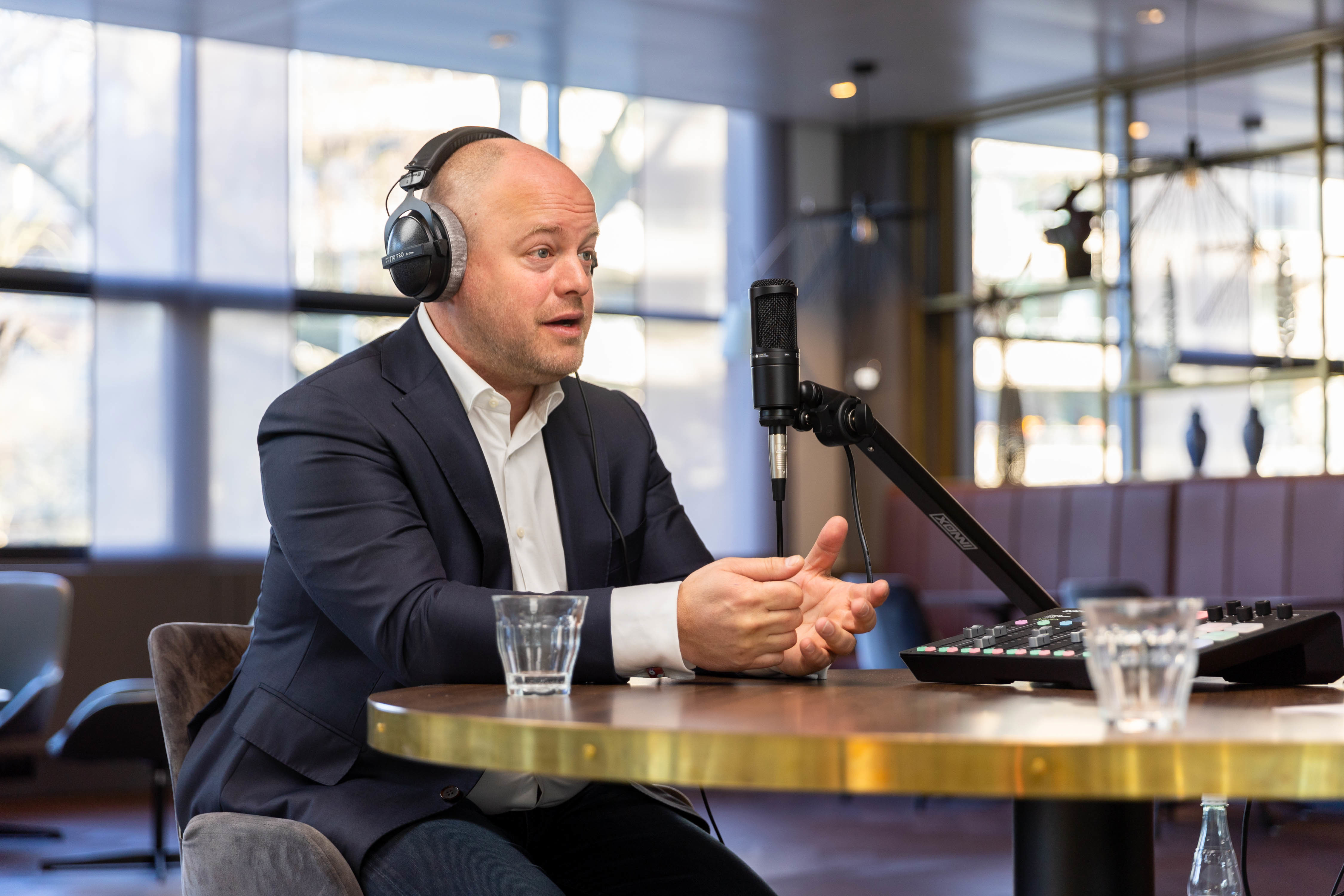 Jochem in een jasje met een koptelefoon zit aan een tafel tijdens een opname, met een microfoon en een mengpaneel voor zich.