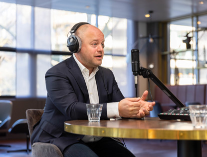 Jochem in een jasje met een koptelefoon zit aan een tafel tijdens een opname, met een microfoon en een mengpaneel voor zich.