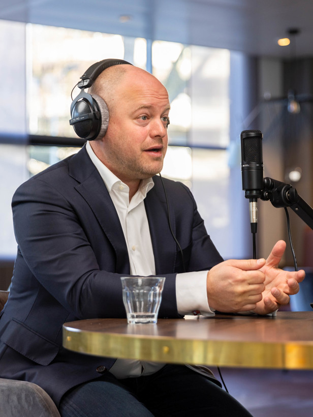 Jochem in een jasje met een koptelefoon zit aan een tafel tijdens een opname, met een microfoon en een mengpaneel voor zich.