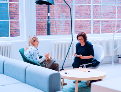 Twee vrouwen zitten in een lichte, moderne ruimte. De ene heeft een notitieboekje en de andere een pen, terwijl ze met elkaar praten.