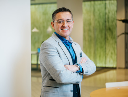 Een man in een lichtgrijze blazer met gekruiste armen staat voor een moderne kantooromgeving met een groene wand.