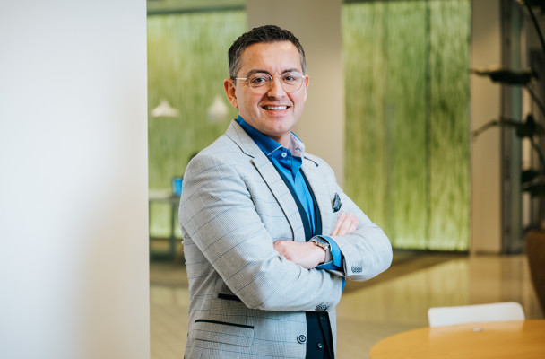 Een man in een lichtgrijze blazer met gekruiste armen staat voor een moderne kantooromgeving met een groene wand.