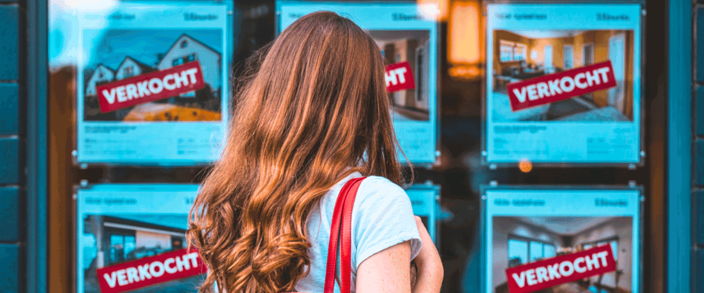 Een vrouw met lang, golvend haar bekijkt woningadvertenties met het grote woord VERKOCHT in het rood op het raam van een makelaarskantoor.