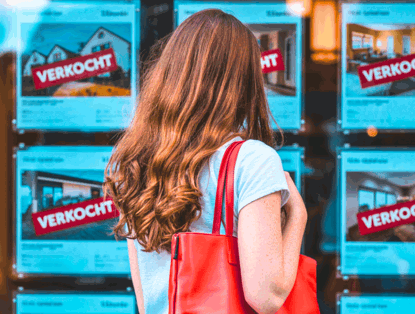 Een vrouw met lang, golvend haar bekijkt woningadvertenties met het grote woord VERKOCHT in het rood op het raam van een makelaarskantoor.