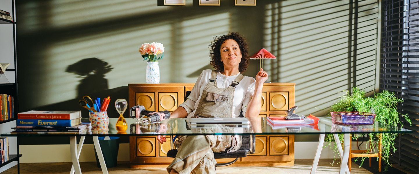 Een persoon zit achter een glazen bureau in een goed verlichte kamer met boekenkasten, een bloempot en kantoorbenodigdheden.