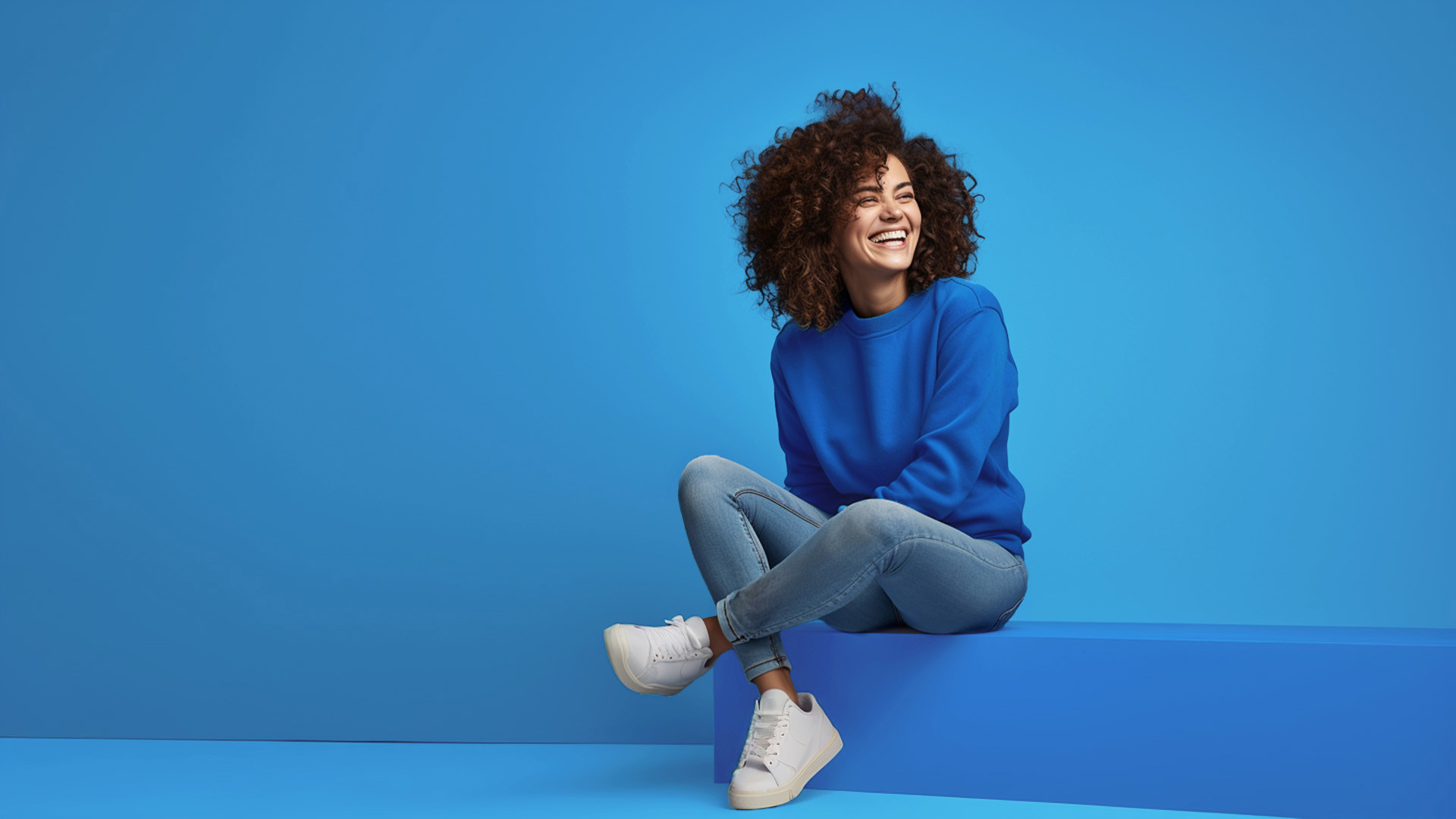 Een vrouw in een blauwe trui zit met gekruiste benen tegen een blauwe achtergrond, in een ontspannen houding.