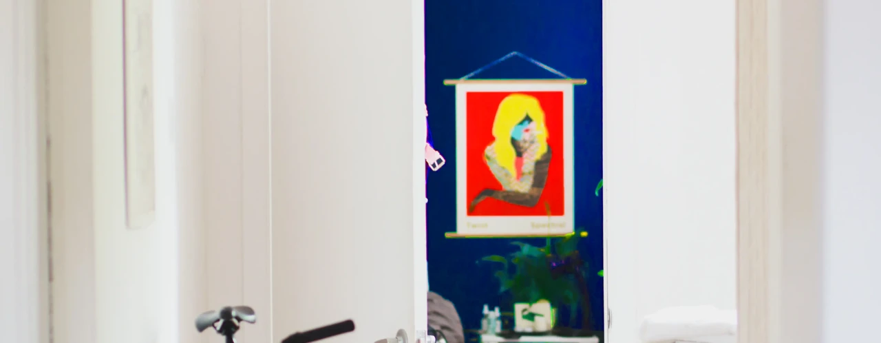 Een kleurrijk kunstwerk van een vrouw in een rode lijst hangt aan de muur, zichtbaar door een open deur.