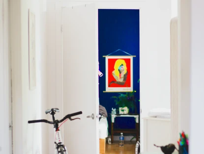 Een kleurrijk kunstwerk van een vrouw in een rode lijst hangt aan de muur, zichtbaar door een open deur.