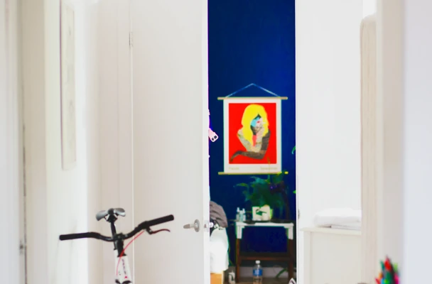 Een kleurrijk kunstwerk van een vrouw in een rode lijst hangt aan de muur, zichtbaar door een open deur.