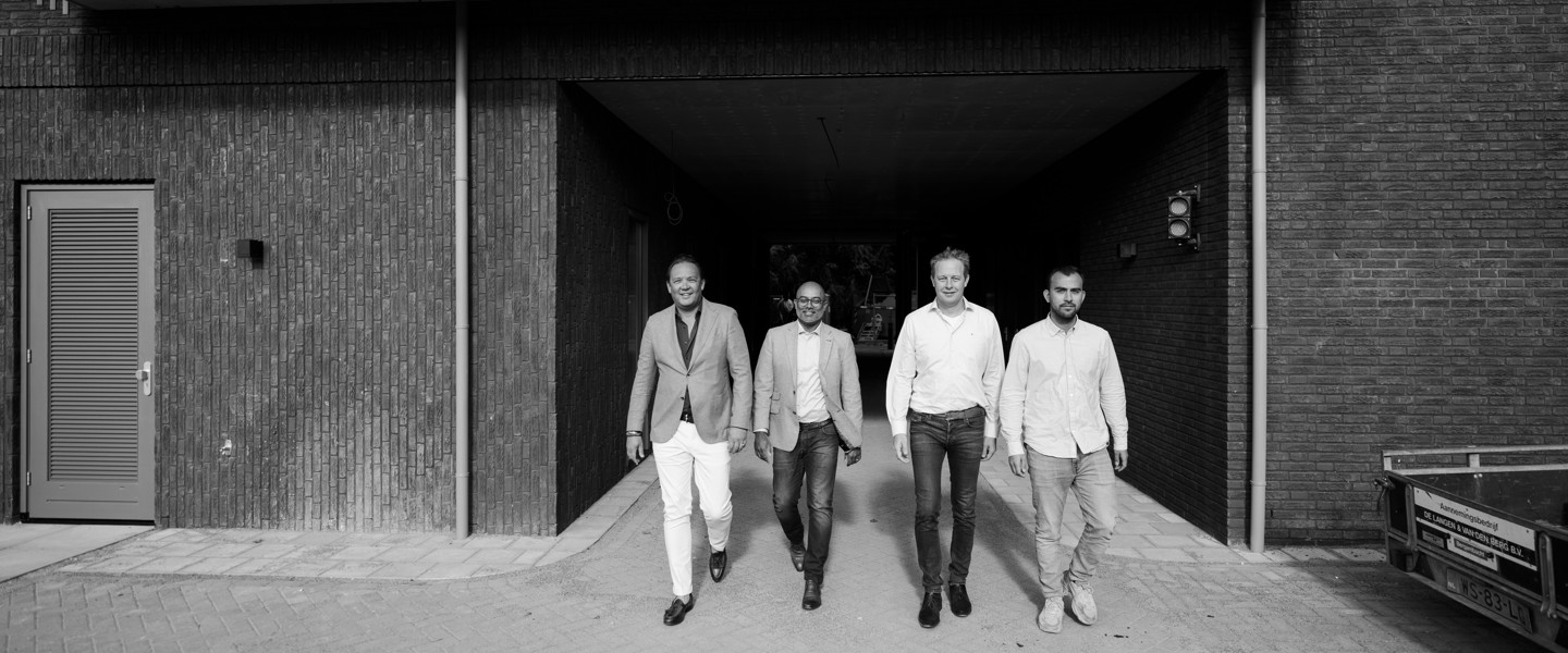 Vier mannen in zakelijke kleding lopen samen uit een donkere gang naar een verlichte buitenruimte.