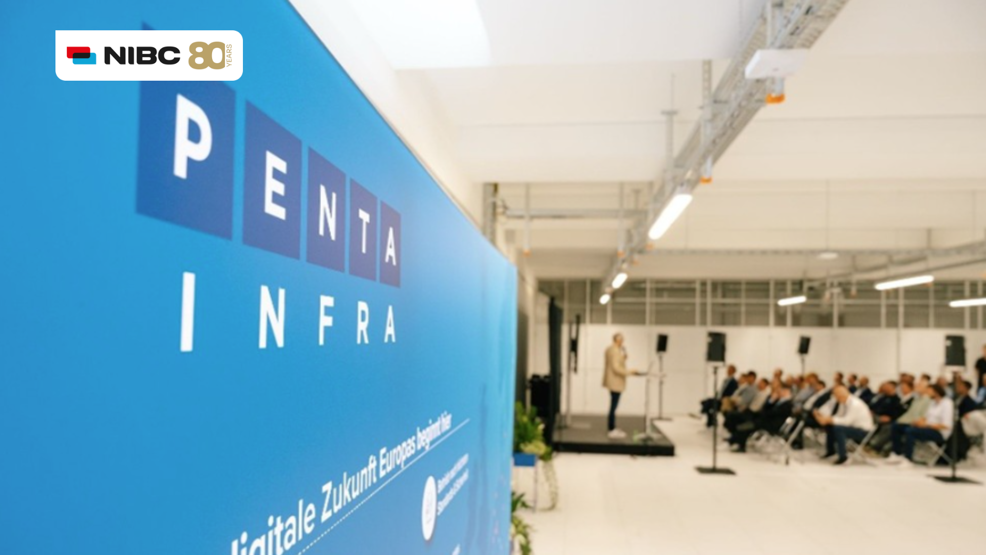 Blauwe muur met de tekst PENTA INFRA en het NIBC-logo, met op de achtergrond een spreker op een podium.