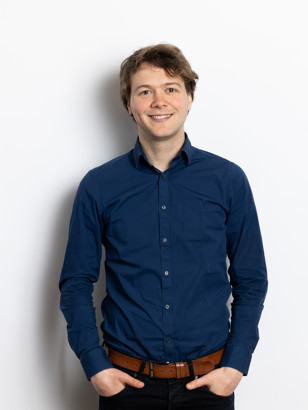 Sjors Van Hooff in een blauw overhemd met een riem, staand voor een witte achtergrond.