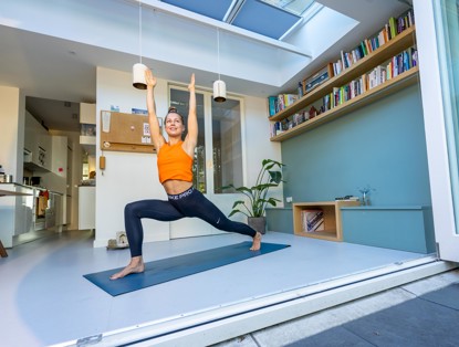 Vrouw in een yogapose in een lichte kamer met planten en boekenplanken, gekleed in een oranje top en zwarte yoga leggings.