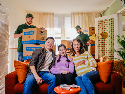 Een gezin zit op een bank in hun woonkamer, omringd door verhuisdozen, terwijl een verhuizer aan de achterkant bezig is.