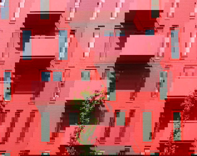 Rood gebouw met grote ramen en twee uitgeronde balkons, omringd door groene planten.