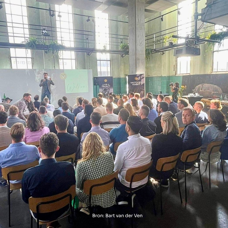 Een groep mensen zit in een zaal en kijkt naar een spreker op het podium, met een projector op de achtergrond.