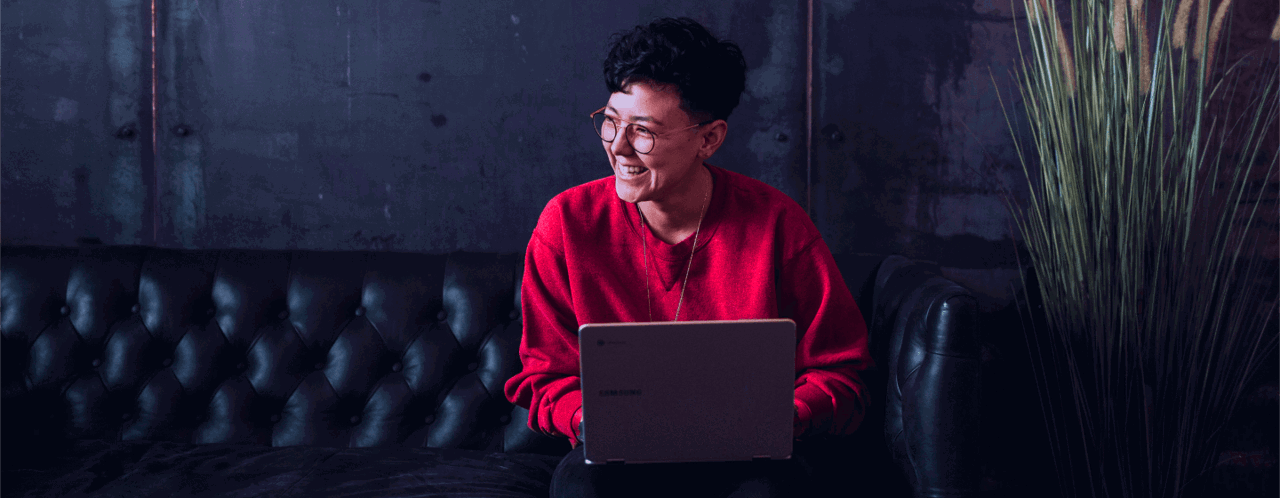 Een persoon in een rode trui zit op een bank met een laptop op schoot, voor een donkere, industriële achtergrond.