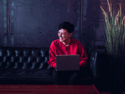 Een persoon in een rode trui zit op een bank met een laptop op schoot, voor een donkere, industriële achtergrond.