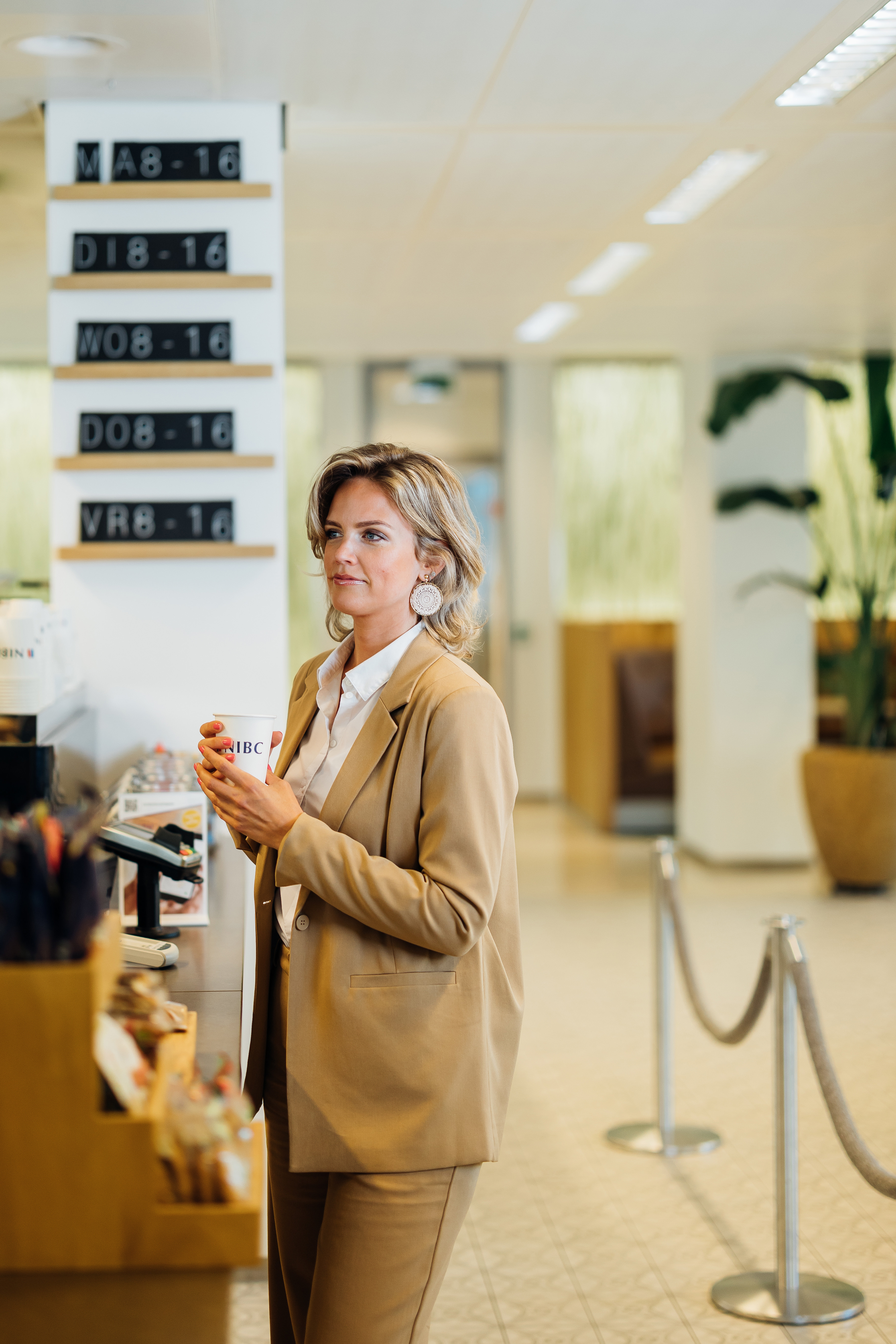 Een vrouw in een beige kostuum staat aan een balie met een koffie in haar hand en kijkt naar de menuborden.