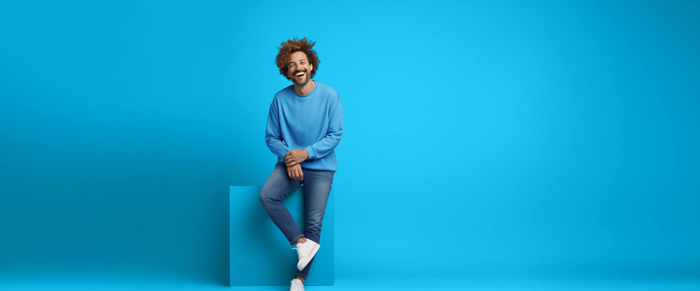 Jongeman in een blauwe trui en jeans leunt relaxed tegen een blauwe achtergrond.