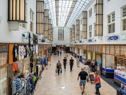 A bustling shopping gallery filled with people shopping, with bright light streaming through large glass skylights.