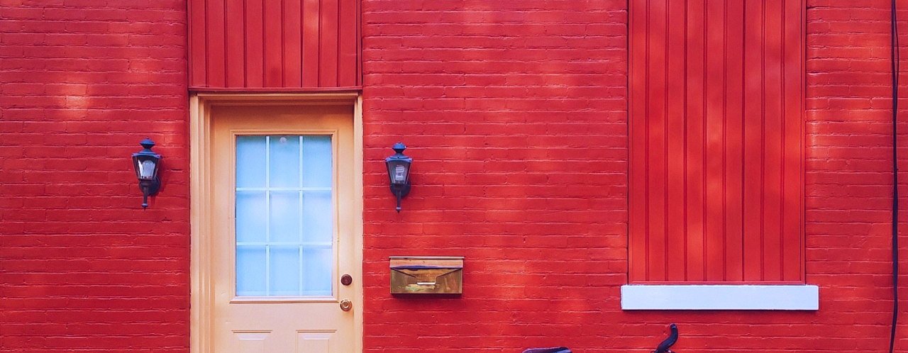 Rode bakstenen muur met een lichte deur en een fiets aan de rechterzijde.
