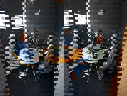 Drie mensen zitten aan een ronde tafel in een opnameset, omringd door microfoons en opnameapparatuur.