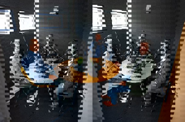 Drie mensen zitten aan een ronde tafel in een opnameset, omringd door microfoons en opnameapparatuur.