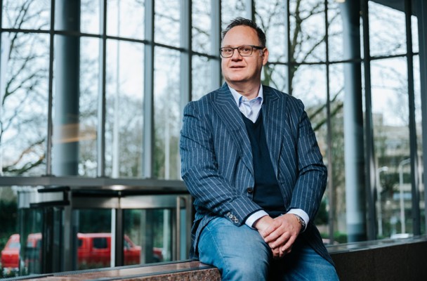 Een man in zakelijke kleding zit op een bank in een lichte, moderne ruimte met grote ramen en een groene achtergrond.