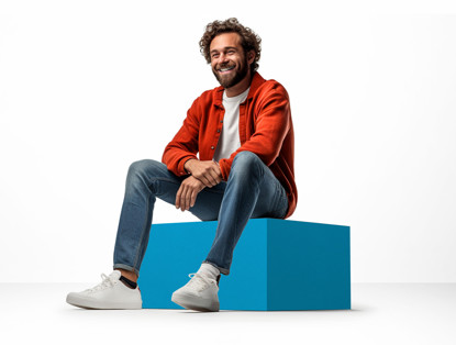 Een persoon in een oranje jas zit ontspannen op een lichtblauwe kubus, gekleed in een wit T-shirt en spijkerbroek.