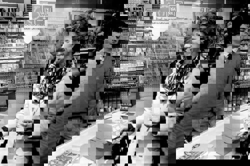 Een vrouw in een winkel staat voor een betaalautomaat met een betaalpas in haar hand. Achter haar zijn producten en een medewerker zichtbaar.