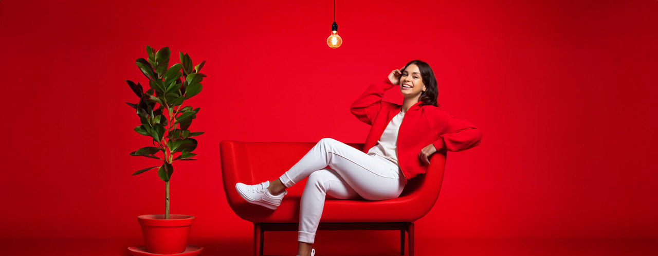 Vrouw in een rode outfit ontspant op een rode bank, met een plant op de voorgrond en een hangende lamp boven haar.