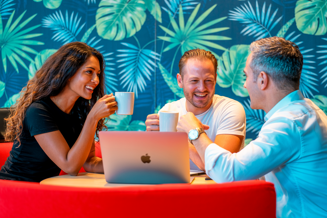 Drie mensen zitten aan een tafel met een laptop en kopjes koffie, omringd door een wand vol planten.