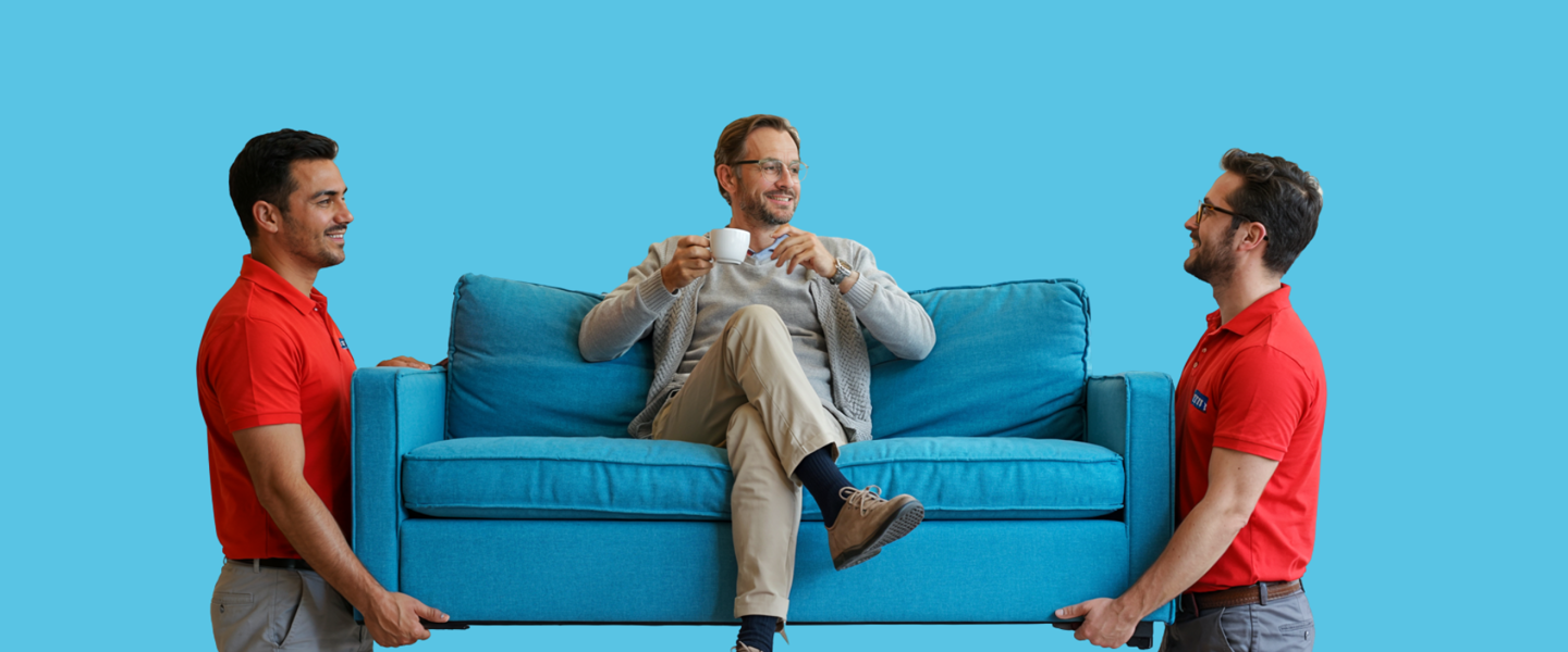 Twee mannen tillen een turquoise bank op, terwijl een derde man op de bank zit en een kopje koffie vasthoudt.