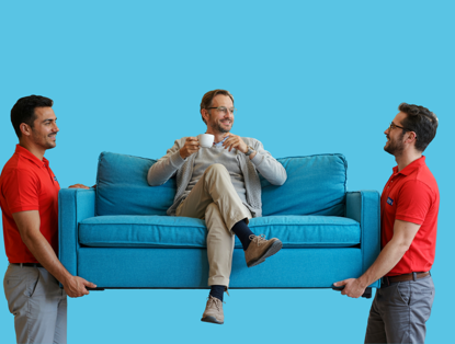 Twee mannen tillen een turquoise bank op, terwijl een derde man op de bank zit en een kopje koffie vasthoudt.