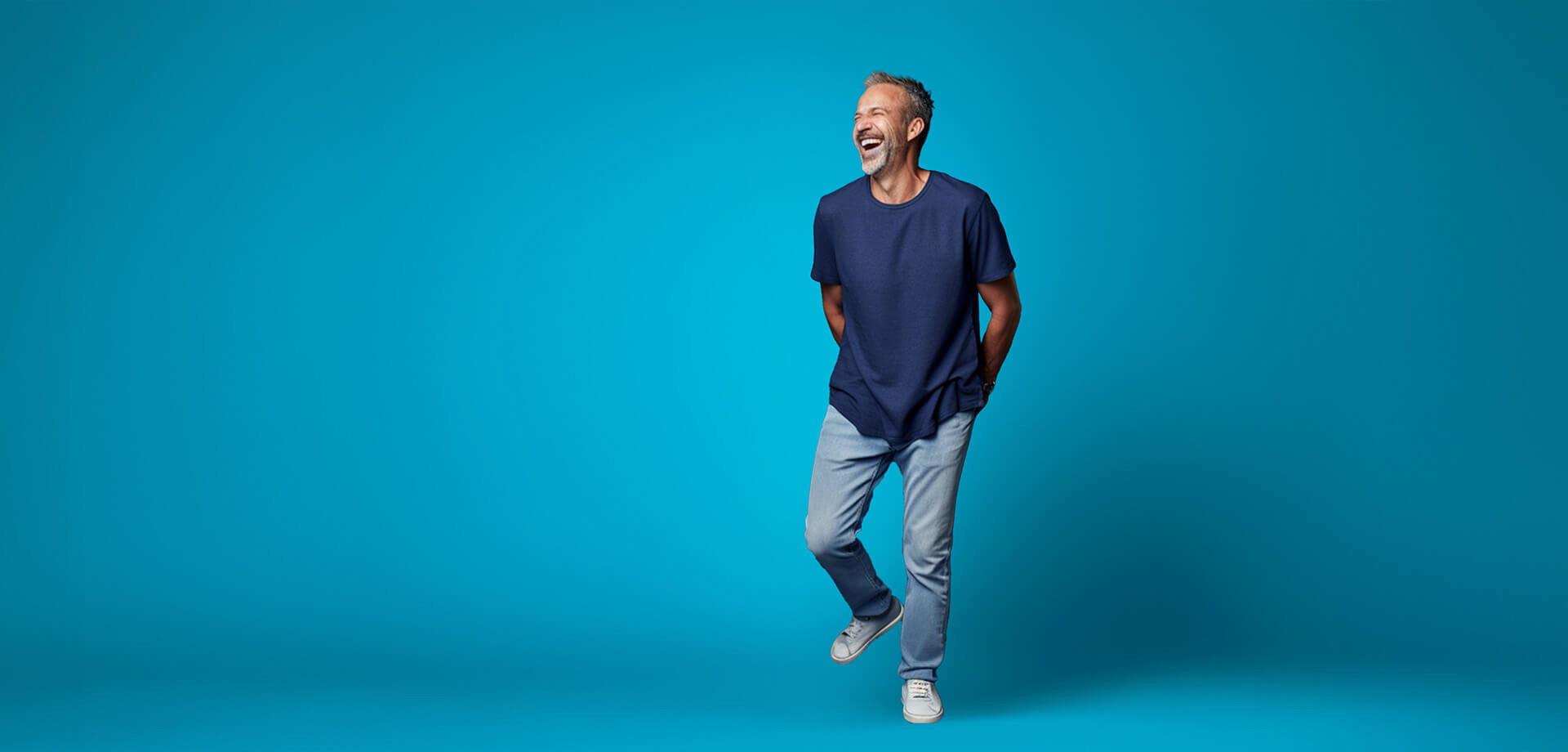 Man in een blauw T-shirt en lichte jeans, staand voor een felblauwe achtergrond.