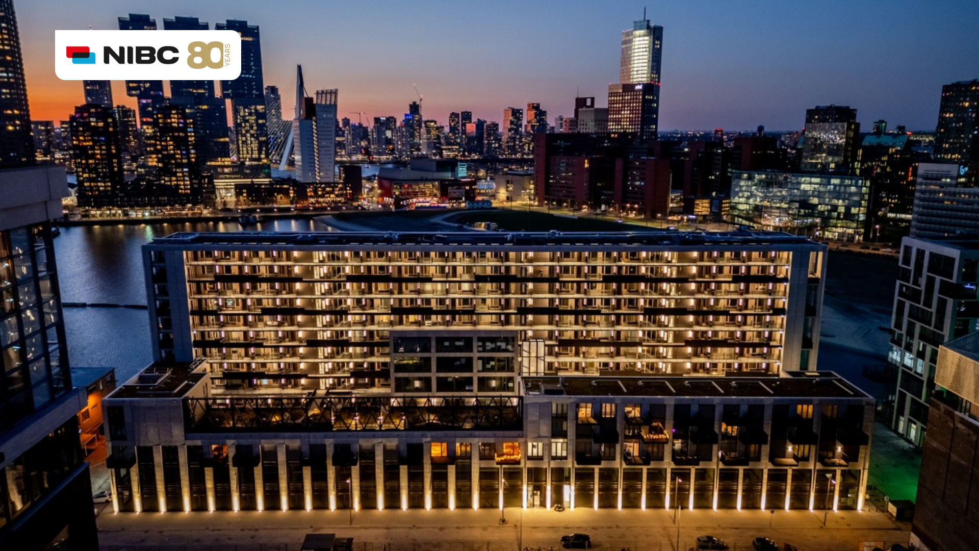 Stadsgezicht van een verlichte skyline met moderne gebouwen en water, met het logo van NIBC 80 jaar bovenaan.