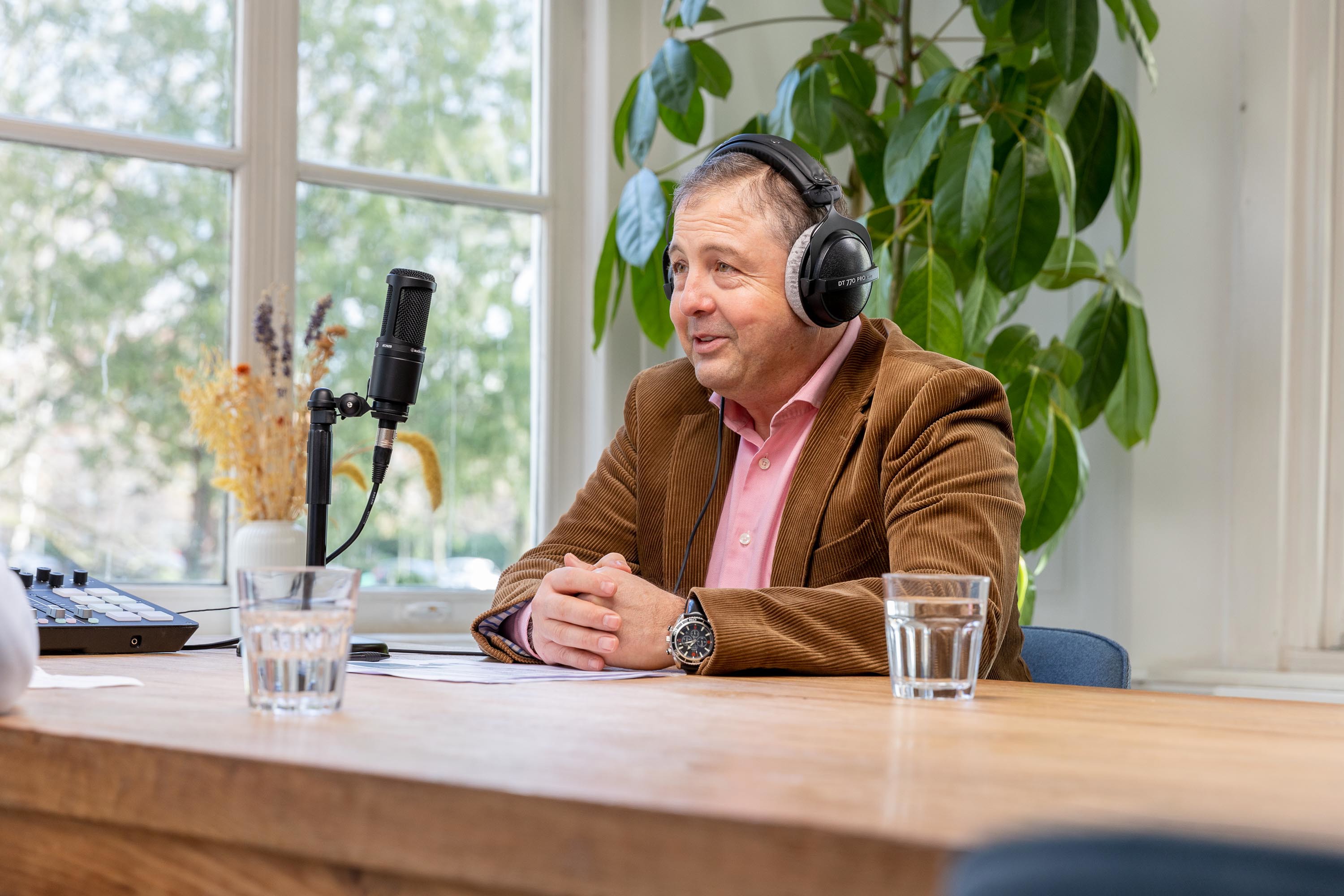 Imed in een bruin jasje met een koptelefoon zit aan een tafel met glazen water en een microfoon, gereed voor een opname.