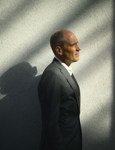 Man in een grijs pak staat zijwaarts tegen een lichte muur, met schaduwpatronen op zijn lichaam.