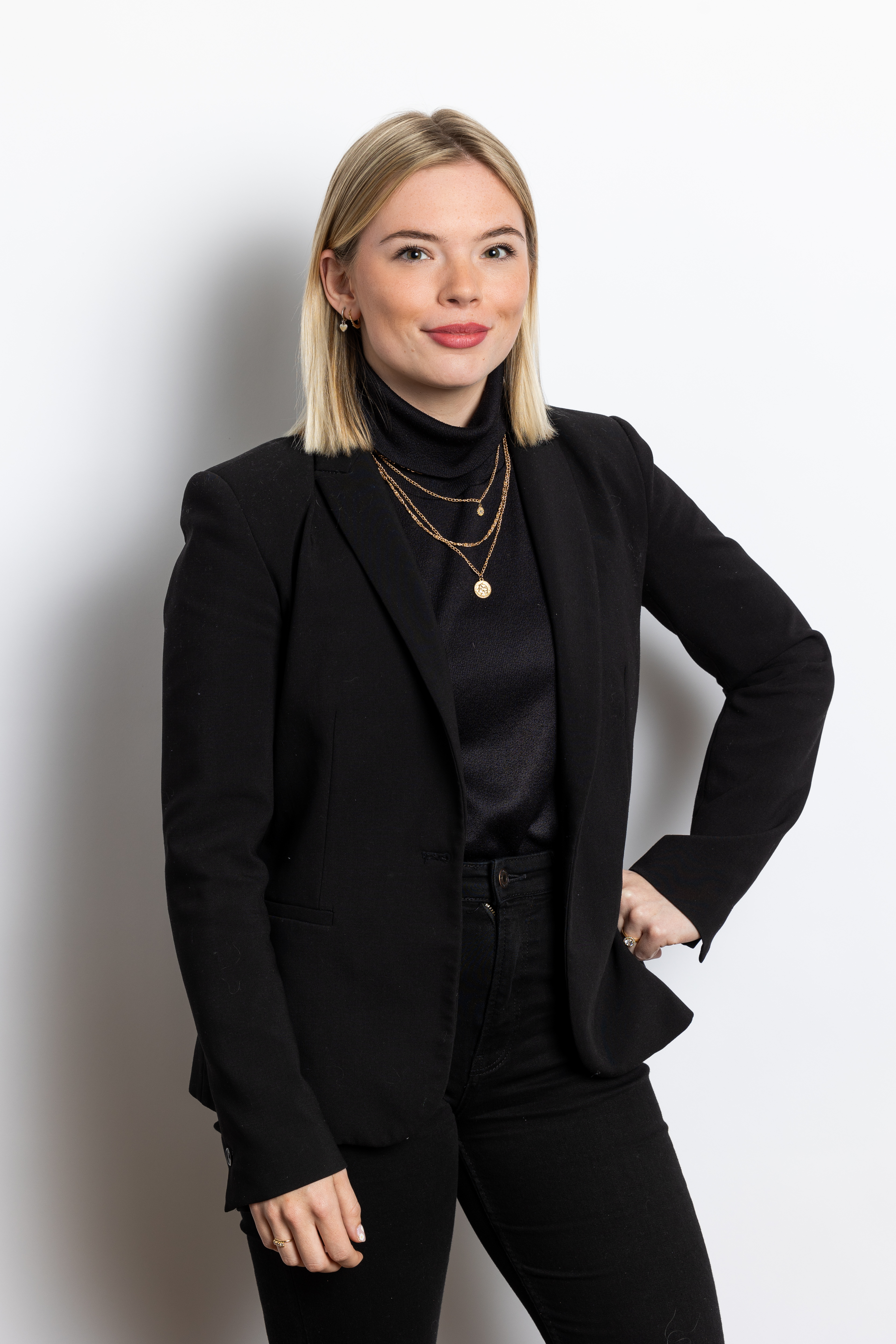Carmen Oogjen in zwarte blazer en top, versierd met meerdere kettingen, poseert met een hand op haar heup tegen een witte achtergrond.