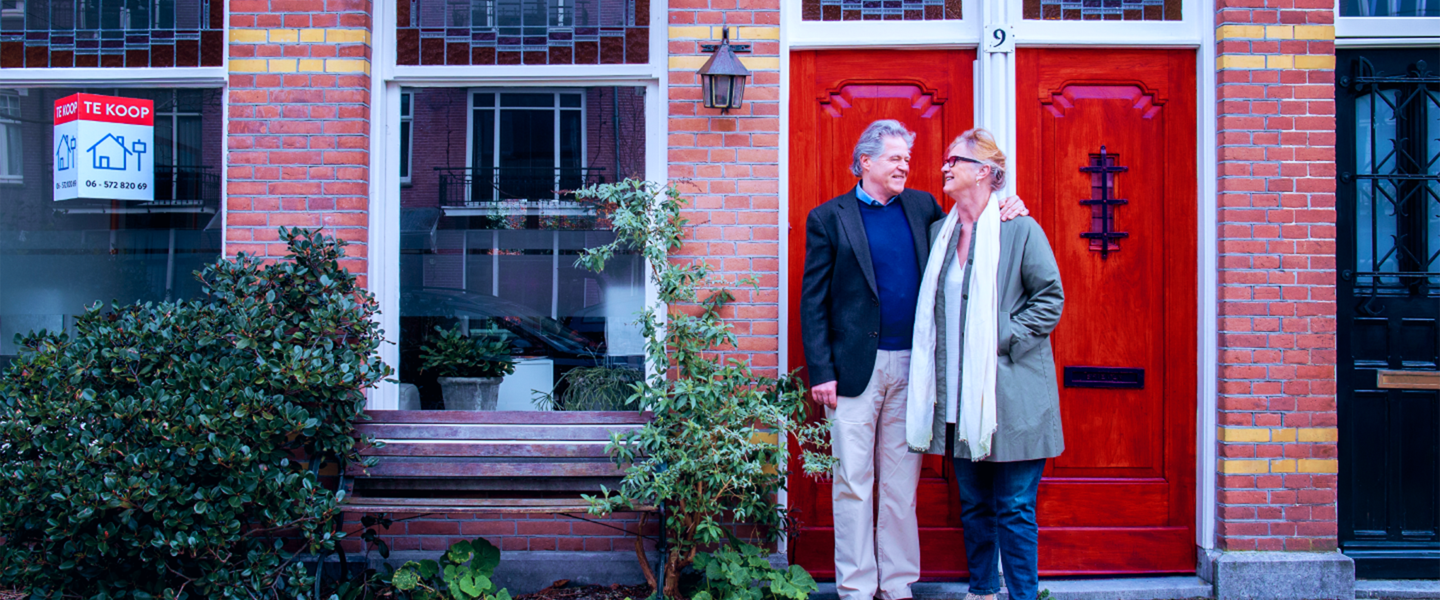 Een man en een vrouw staan naast een huis met een 'Te koop'-bord bij de deur, omgeven door groen.