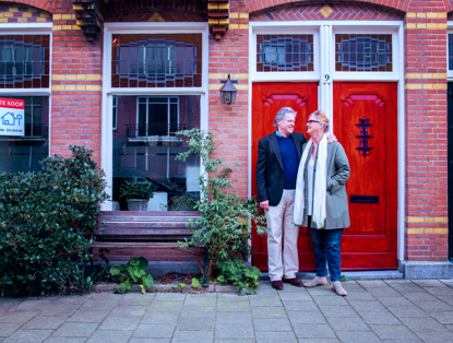 Een man en een vrouw staan naast een huis met een 'Te koop'-bord bij de deur, omgeven door groen.