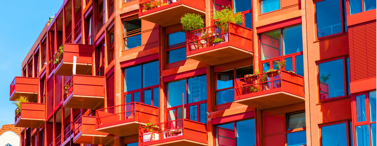 Modern red apartments with glass balconies, each surrounded by greenery.