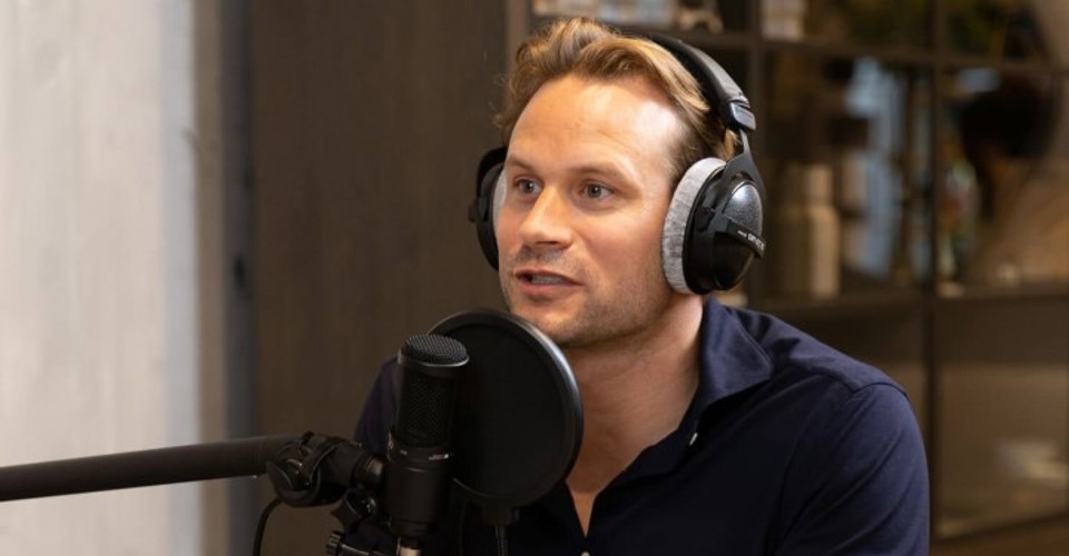 Dirk Taat in een zwart overhemd met een hoofdtelefoon zit voor een microfoon in een moderne studio, terwijl hij geïnterviewd wordt.