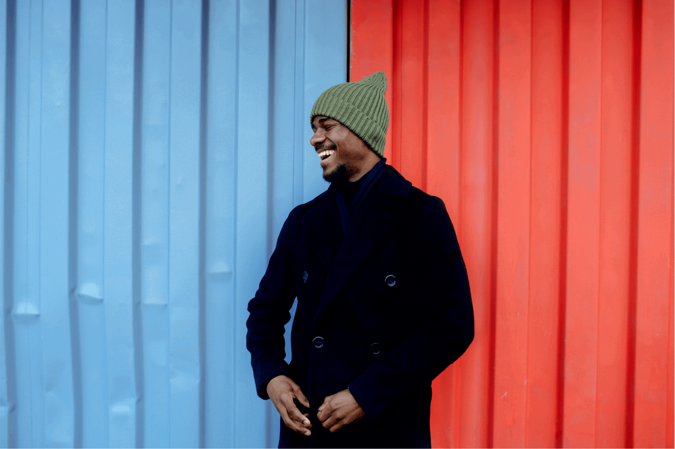 Man in een zwarte winterjas met een groene muts, staand tussen een blauwe en een rode achtergrond.