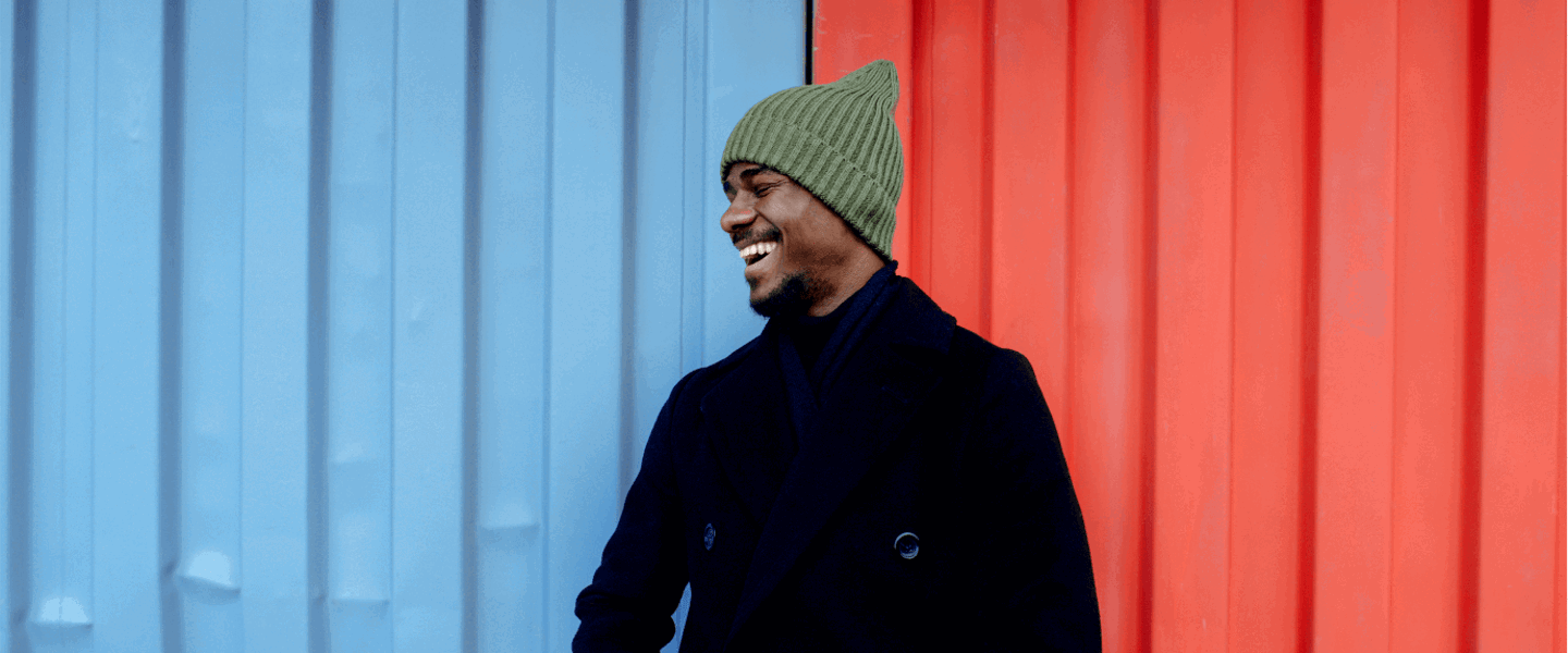 Man in een zwarte winterjas met een groene muts, staand tussen een blauwe en een rode achtergrond.