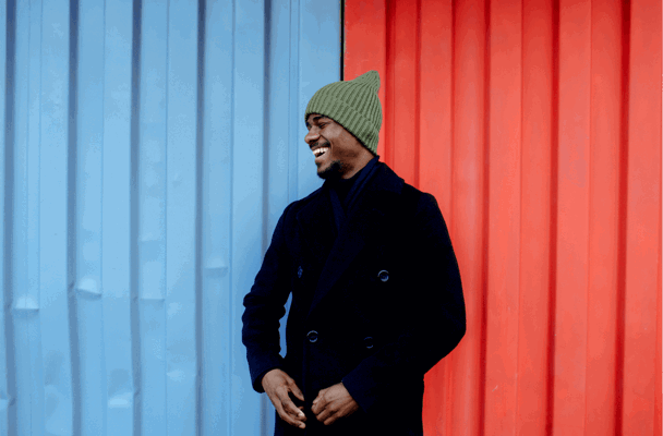 Man in een zwarte winterjas met een groene muts, staand tussen een blauwe en een rode achtergrond.